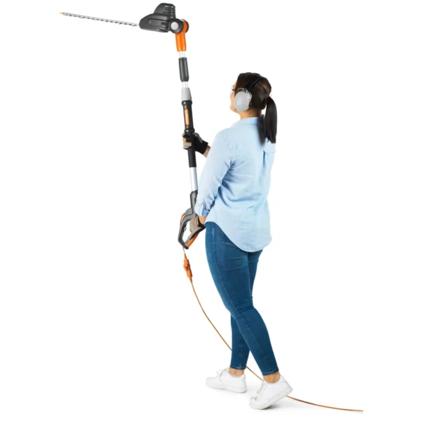 Woman in jeans and a light shirt holds a long extendable hedge trimmer, wearing gloves and earmuffs against a white background.